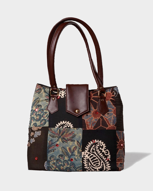 A professional studio shot of the Handcrafted Patchwork Tote Bag. The bag features a sophisticated dark-toned patchwork of floral, paisley, and leaf patterns, accented with dark brown faux leather handles and a magnetic snap closure flap.