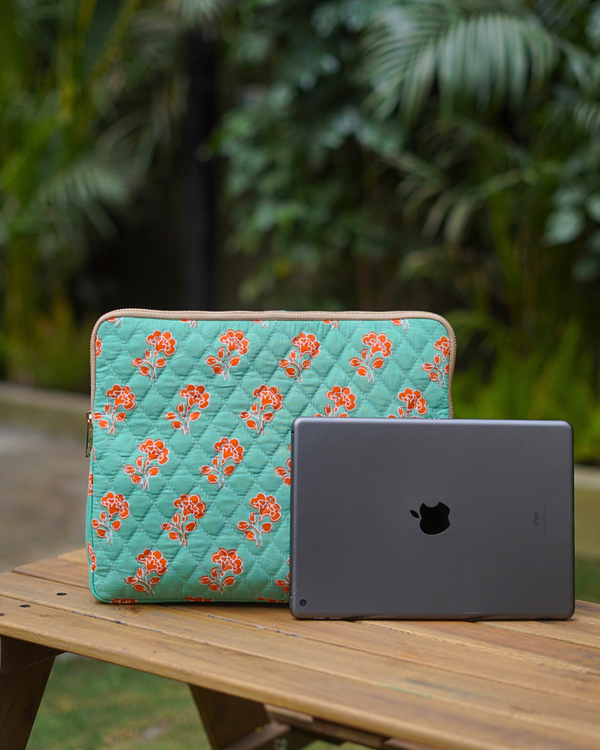 A lifestyle shot of the Leaf & Tile Mosaic Laptop Bag resting on a wooden outdoor table next to a slate-grey laptop, showcasing the vibrant mint green fabric with orange floral and diamond quilt patterns.