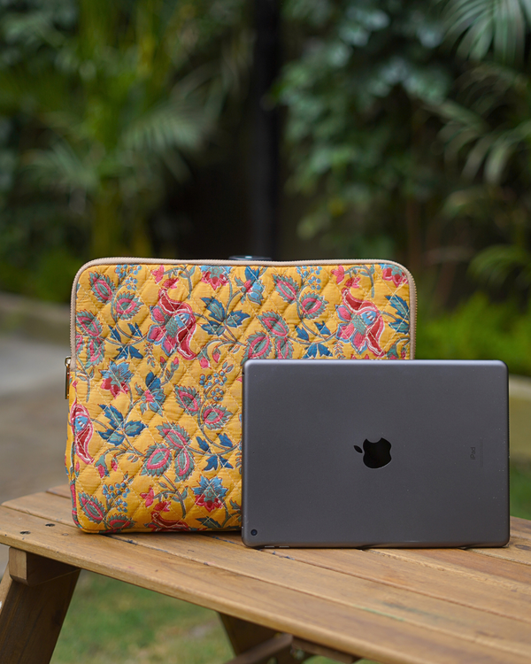 A vibrant lifestyle shot of the Yellow Vine & Blossom Laptop Bag on a wooden outdoor table, positioned next to a slate-grey laptop to highlight its bright yellow base and intricate botanical block-print.