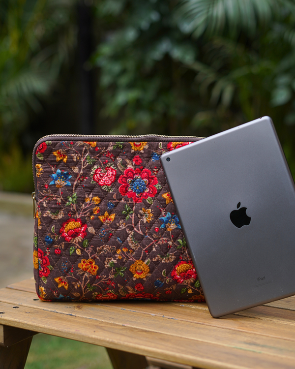 A lifestyle photograph of the Bouquet & Earth-Tone Brown Laptop Bag sitting on a wooden outdoor table next to a slate-grey laptop, highlighting the rich brown quilted fabric and vibrant floral bouquet block-print.