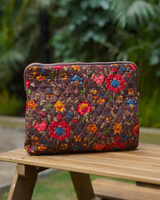 A front-facing shot of the laptop bag standing upright on a wooden surface, set against a lush, green natural background to emphasize the colorful red, blue, and yellow floral details.