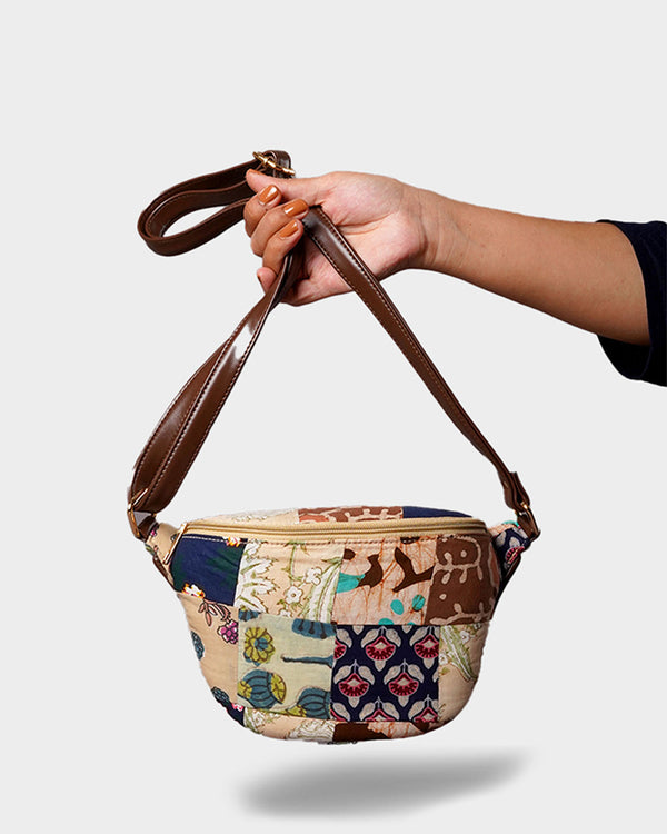 A hand holding the Patchwork Convertible Sling Bag by its dark brown faux leather strap. The bag features an earthy-toned patchwork design with intricate floral, geometric, and leaf patterns.