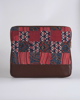 A handcrafted quilted laptop bag featuring a classic Ajrakh-inspired patchwork design in crimson red and deep indigo blue. The panels showcase traditional floral block prints and geometric motifs, set above a smooth, dark chocolate-brown base.