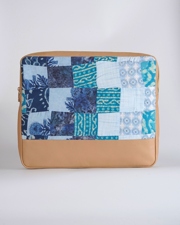 A handcrafted quilted laptop bag featuring a coastal-inspired patchwork of indigo, azure, and sky blue. The design highlights traditional batik textures, wave patterns, and delicate floral block prints, finished with a smooth tan-colored base.