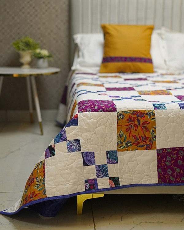 A lifestyle shot of the Modern Mosaic Patchwork Quilt on a bed, showing the intricate grid design and vibrant floral blocks against a white fluted headboard.