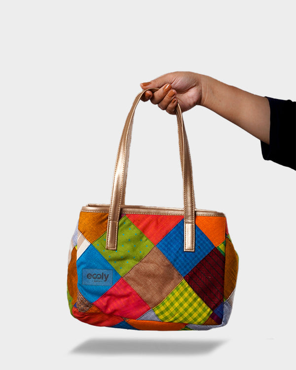 A hand holding the Vibrant Multi-Color Patchwork Handbag by its metallic gold handles. The bag features a colorful geometric patchwork of blue, orange, brown, and green.