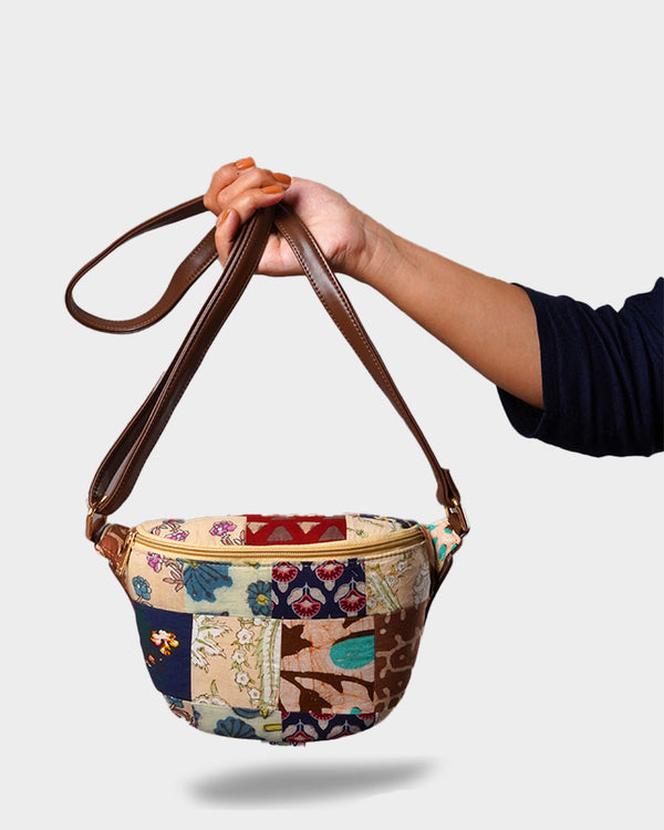 A hand holding the Bohemian Patchwork Convertible Sling Bag by its dark brown adjustable strap. The bag features a vibrant, multi-colored patchwork design with floral and geometric patterns in navy, burgundy, and cream.