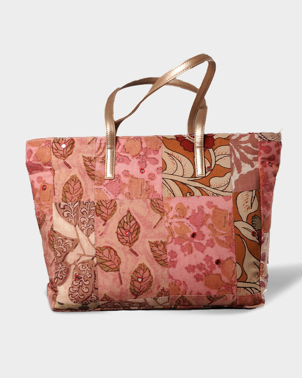 A studio shot of the Spacious Quilted Tote Bag, showcasing a warm patchwork of dusty rose, coral, and chocolate brown floral and leaf patterns with metallic gold shoulder handles.