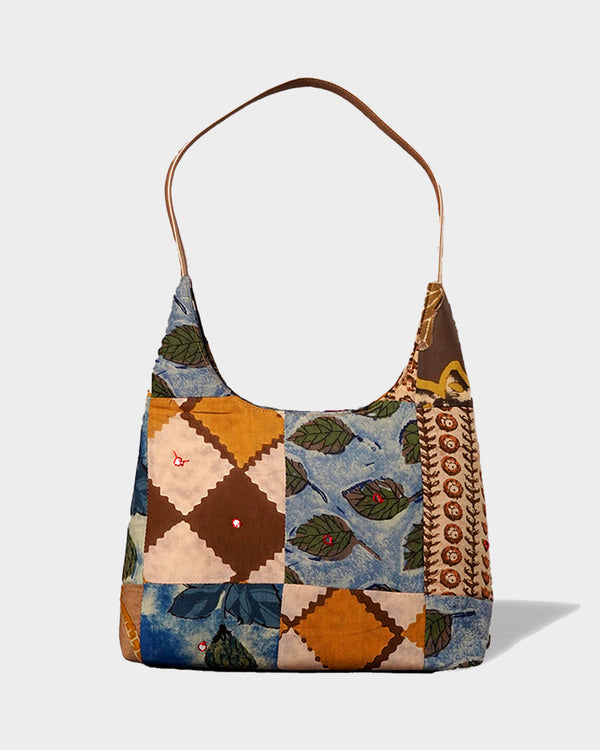 A studio shot of the Earth Tones Patchwork Shoulder Bag, showing its tall hobo silhouette and intricate patchwork of diamond tiles, paisley, and floral motifs in shades of ochre, navy, and brown.