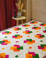A top-down view of the quilt on the bed, highlighting the repeating geometric patchwork of orange, red, and green squares against the neutral beige field.