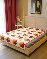 A bright bedroom scene featuring the Red & Beige Patchwork Cotton Bedquilt spread over a bed with a beige upholstered headboard, styled with a small side table and floral decor.