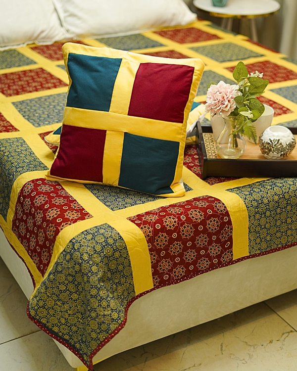 A lifestyle shot of a bed styled with the Bold Retro Checkerboard Quilted Bedspread and matching shams, featuring a large square pillow with a bold yellow cross-pattern over teal and red blocks.